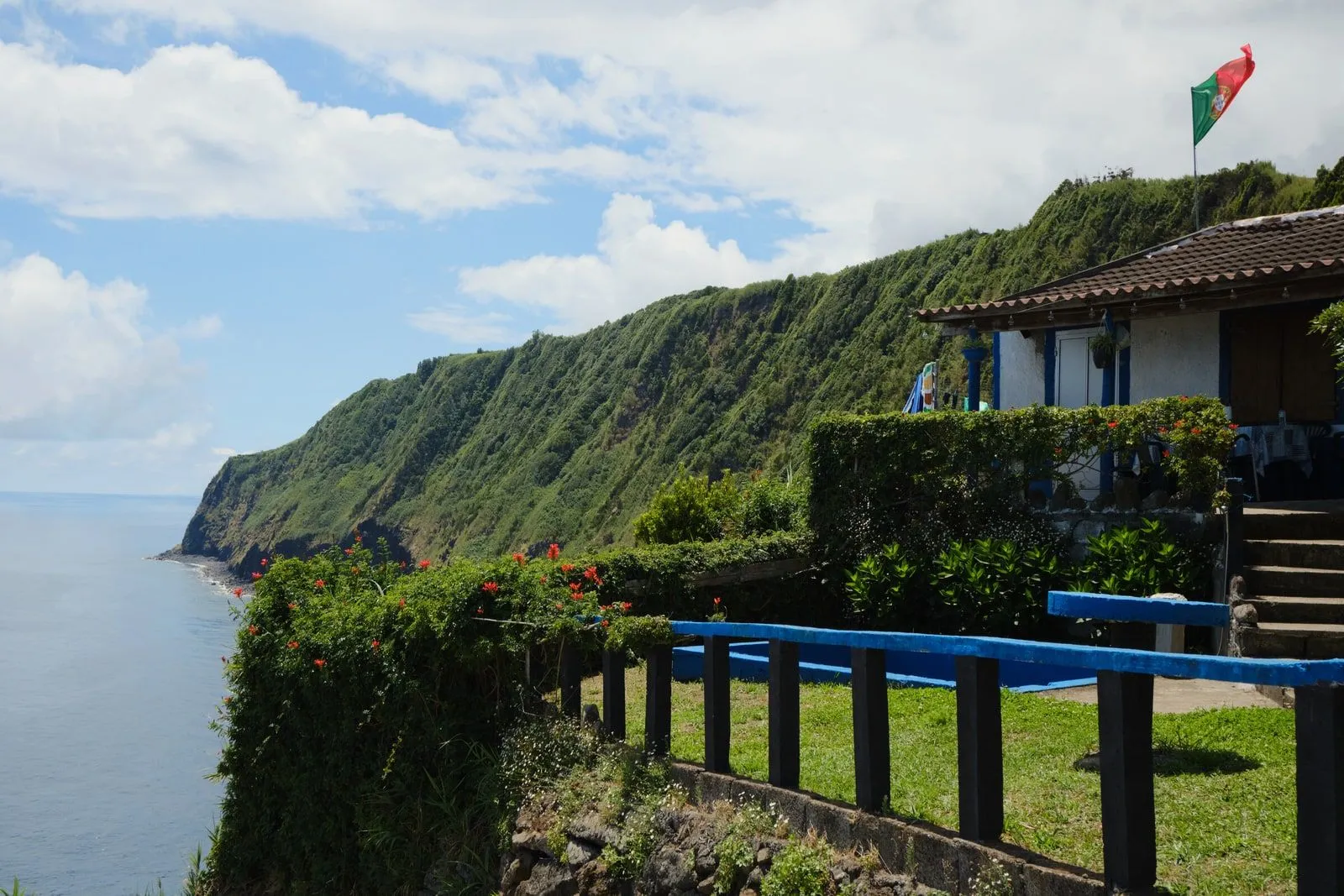 A small house perched above a green cliffside, bay visible beyond.