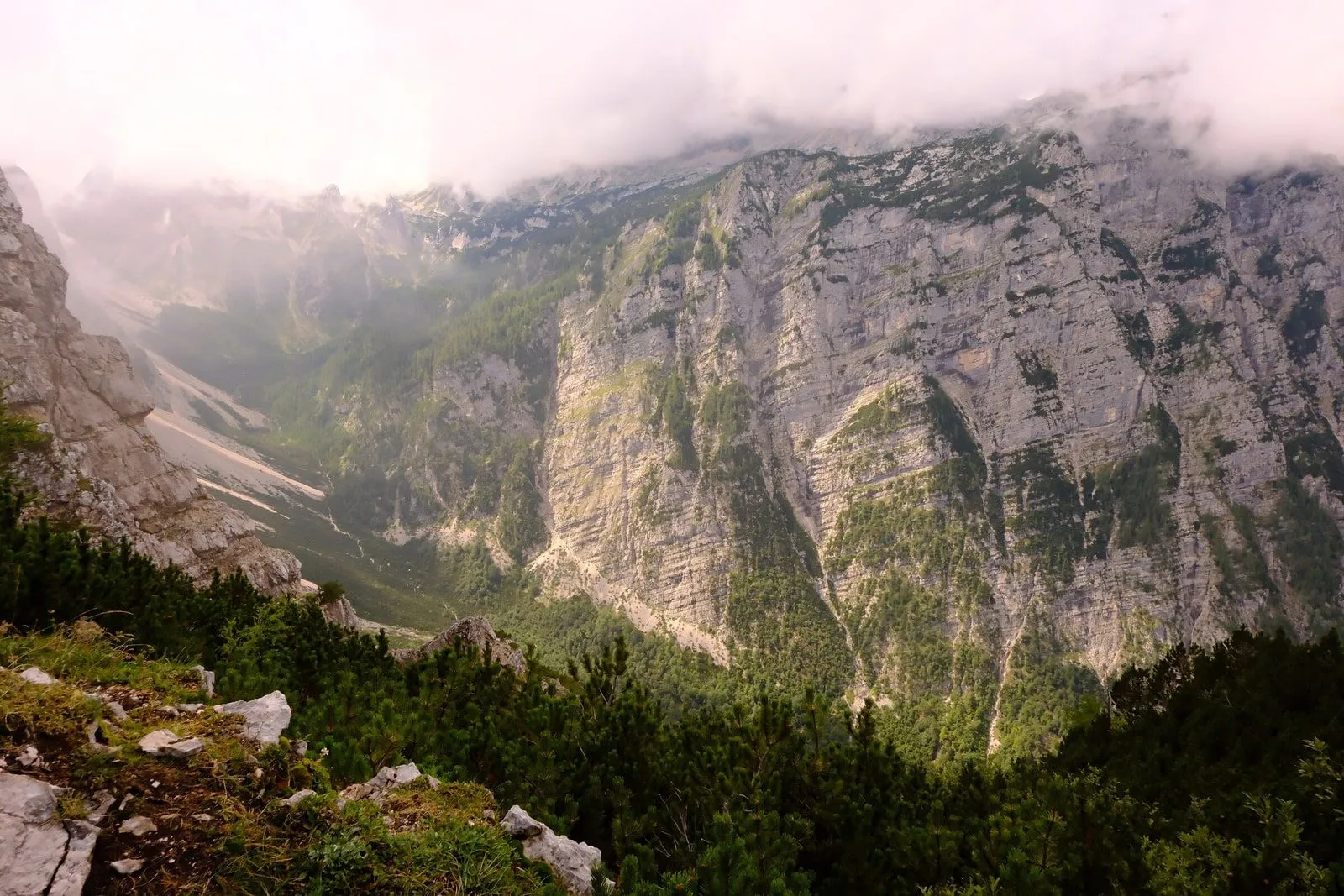 A sheer limestone cliff face above a green alpine valley, cloud clinging to the peak.